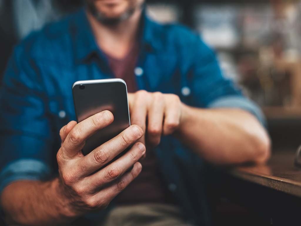 Muškarac koji sjedi za stolom usredotočen je na svoj mobitel, angažiran u razgovoru ili pregledavanju sadržaja