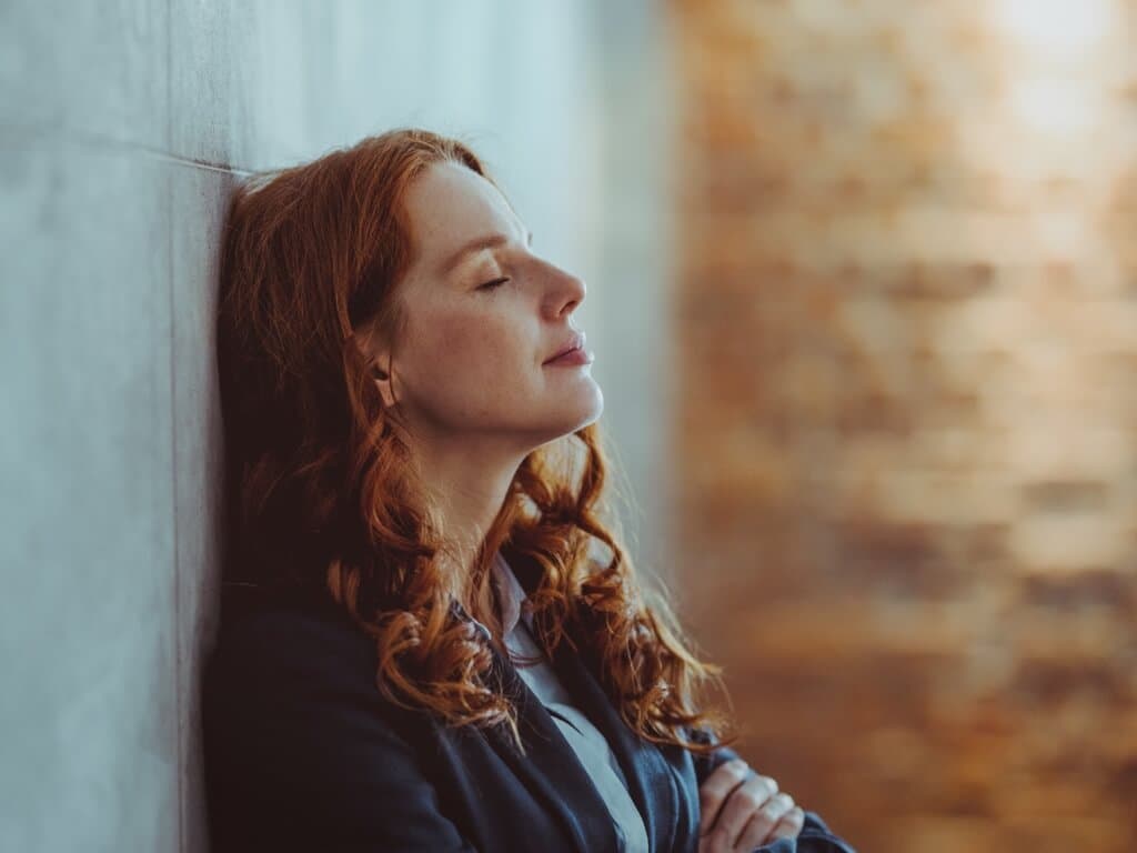 Žena zatvorenih očiju naslanja se na zid, djelujući zamišljeno i spokojno u svom držanju
