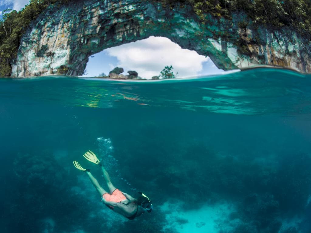 Muškarac ronilački diše u vodi pokraj slikovitog luka, okružen bistrim plavim morem i jarkim sunčevim svjetlom