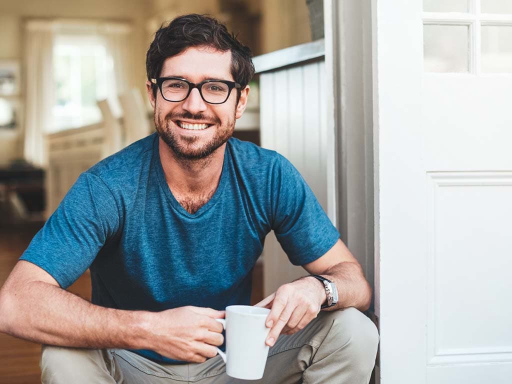 Muškarac s naočalama i plavom košuljom sjedi na podu, držeći šalicu kave u rukama