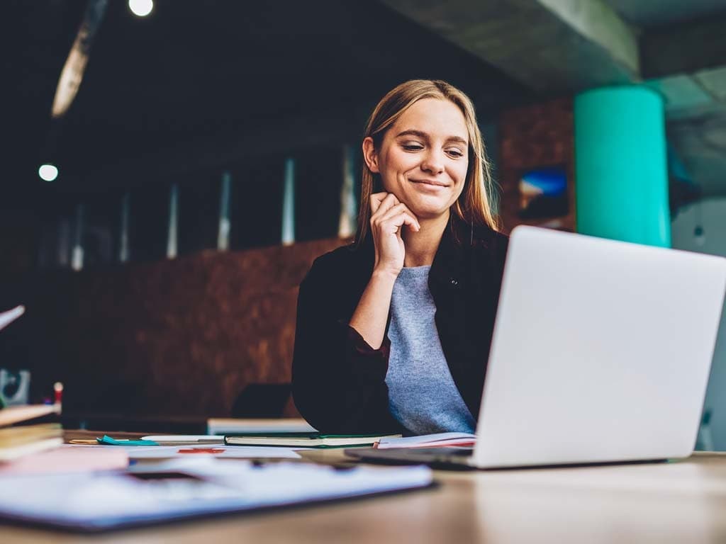 Žena sjedi za stolom, usredotočena na svoj laptop, s bilježnicom i olovkom pored sebe