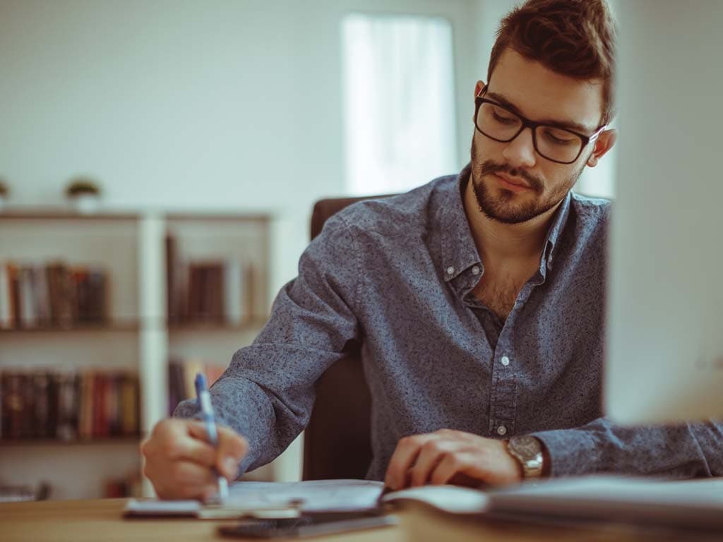 Muškarac s naočalama usredotočeno piše na komadu papira za stolom