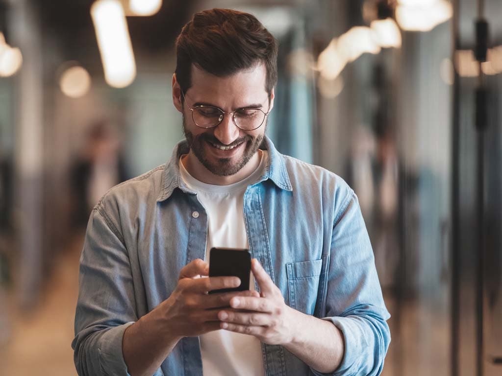 Muškarac u plavoj košulji smiješi se dok gleda u svoj telefon, izgledajući angažirano i sretno