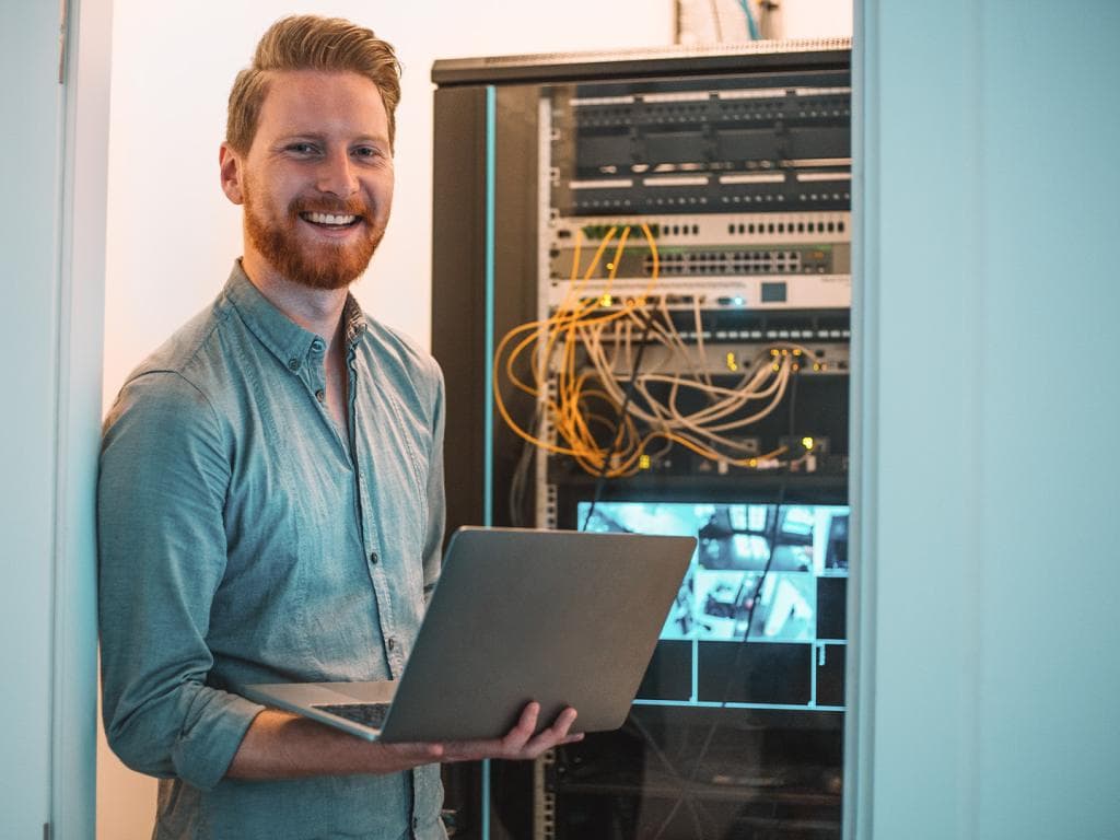 Muškarac stoji s prijenosnim računalom ispred poslužitelja, prikazujući radno okruženje usmjereno na tehnologiju