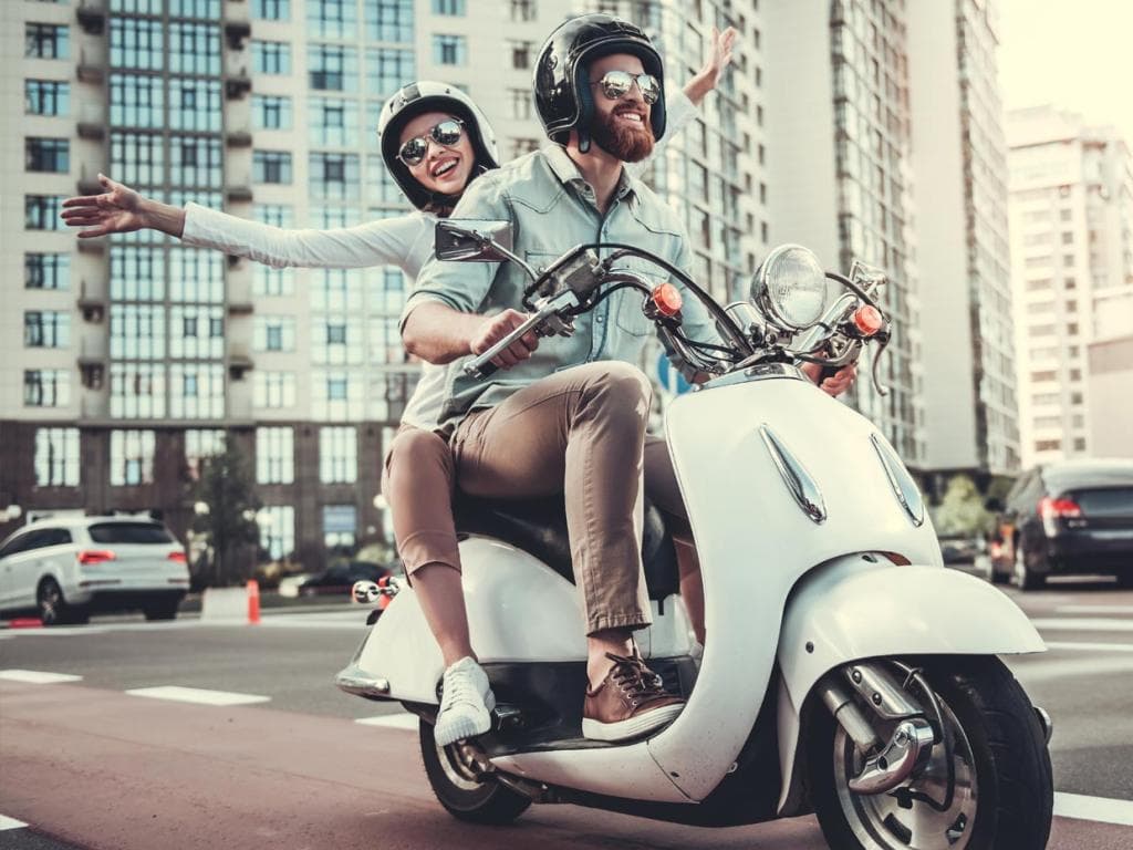Par uživa u vožnji skuterom kroz živopisni grad, s gradskim prizorima i ljudima u pozadini