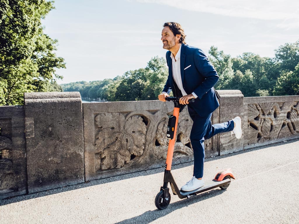 Muškarac u odijelu vozi električni romobil gradskom ulicom, prikazujući spoj profesionalizma i moderne mobilnosti